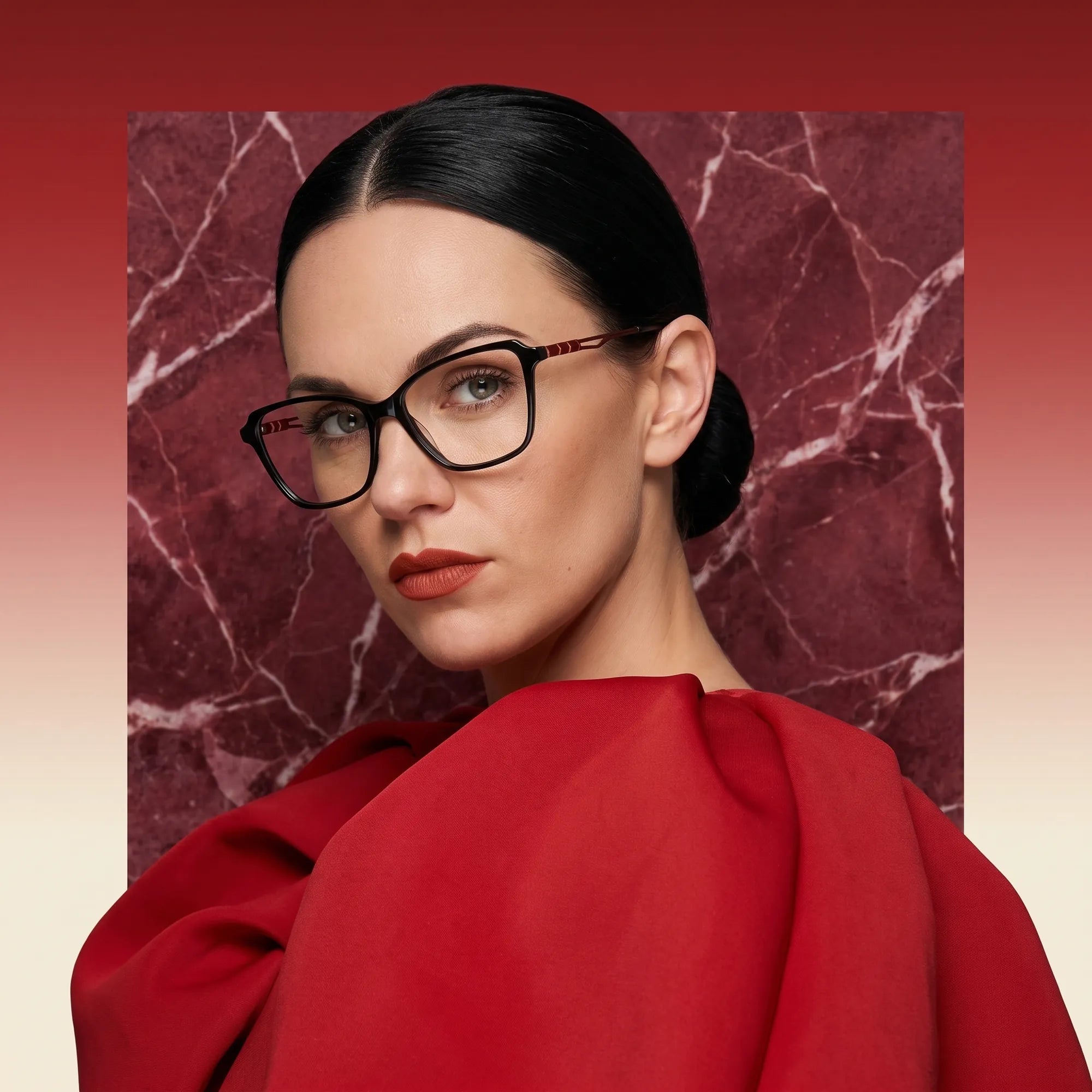 Fiore 1006 eyeglasses in black, worn by a model – campaign portrait, red marble backdrop and draped scarlet dress