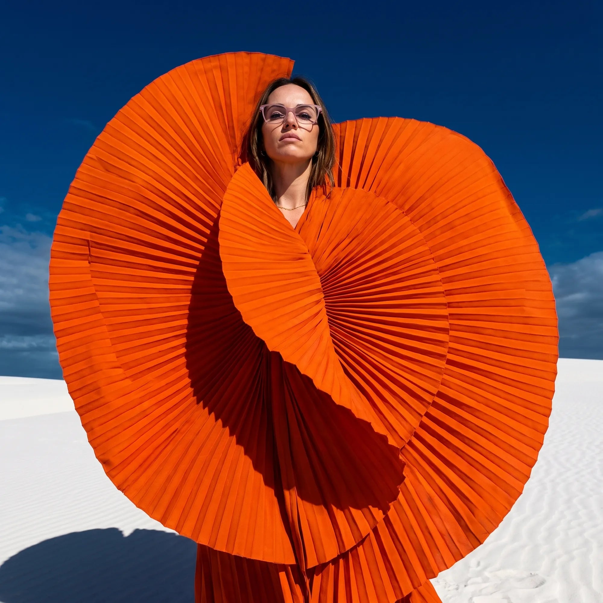 Fiore 1006 eyeglasses in pink, worn by a model – campaign portrait, sculptural orange pleated fan on white dunes
