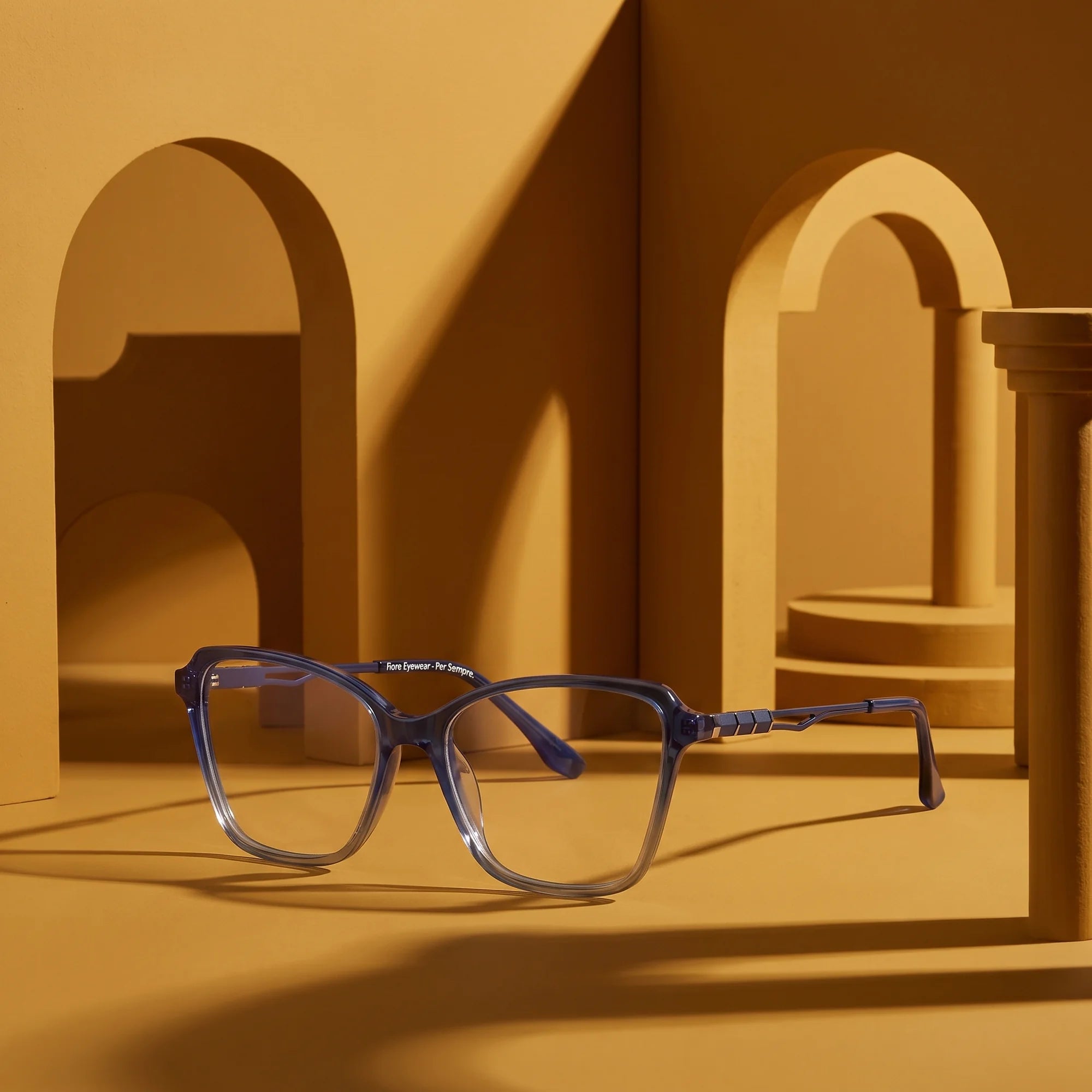 Fiore 1006 eyeglasses in blue – displayed on a mustard ochre podium among arches