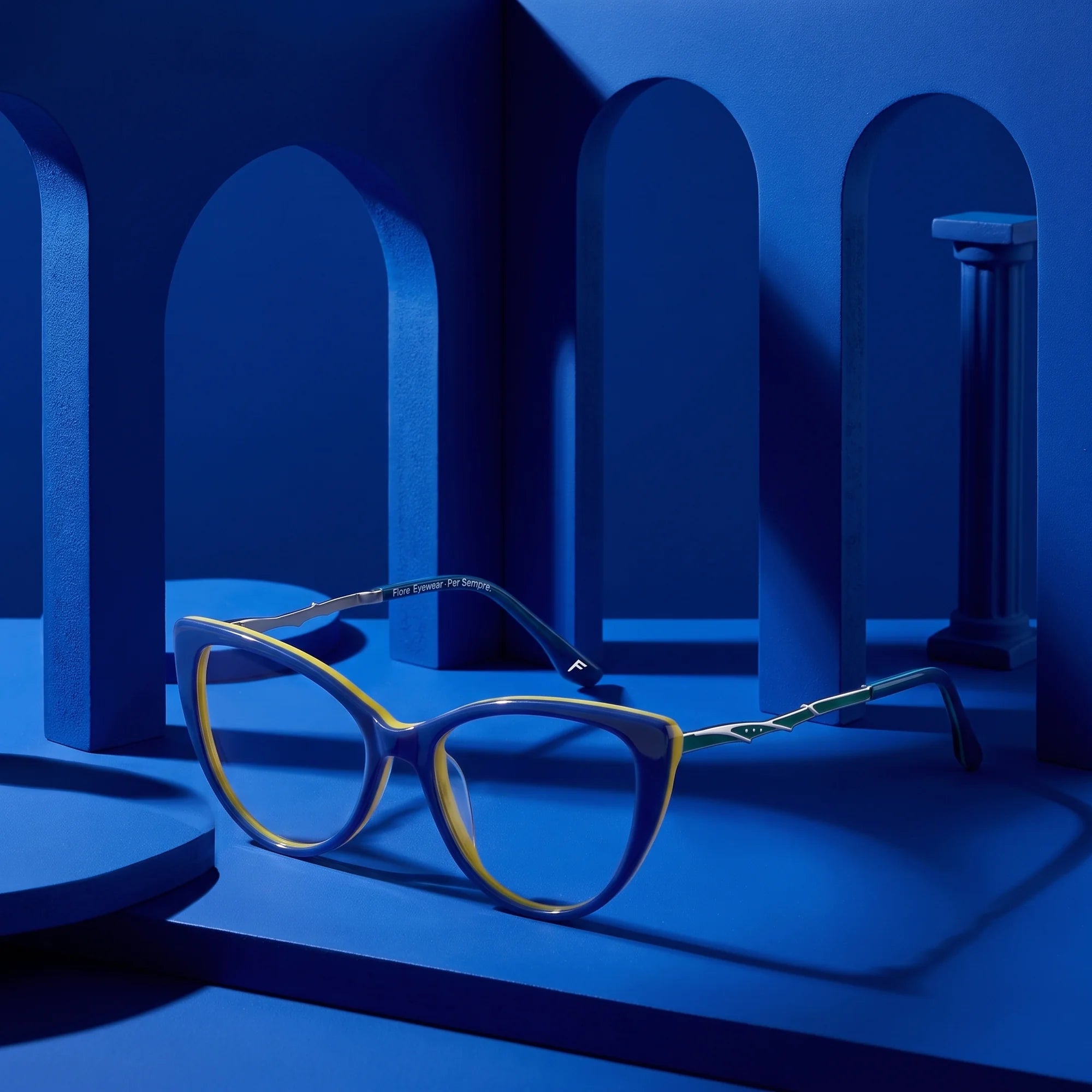 Fiore 1010 eyeglasses in blue green – displayed on a cobalt podium among arches