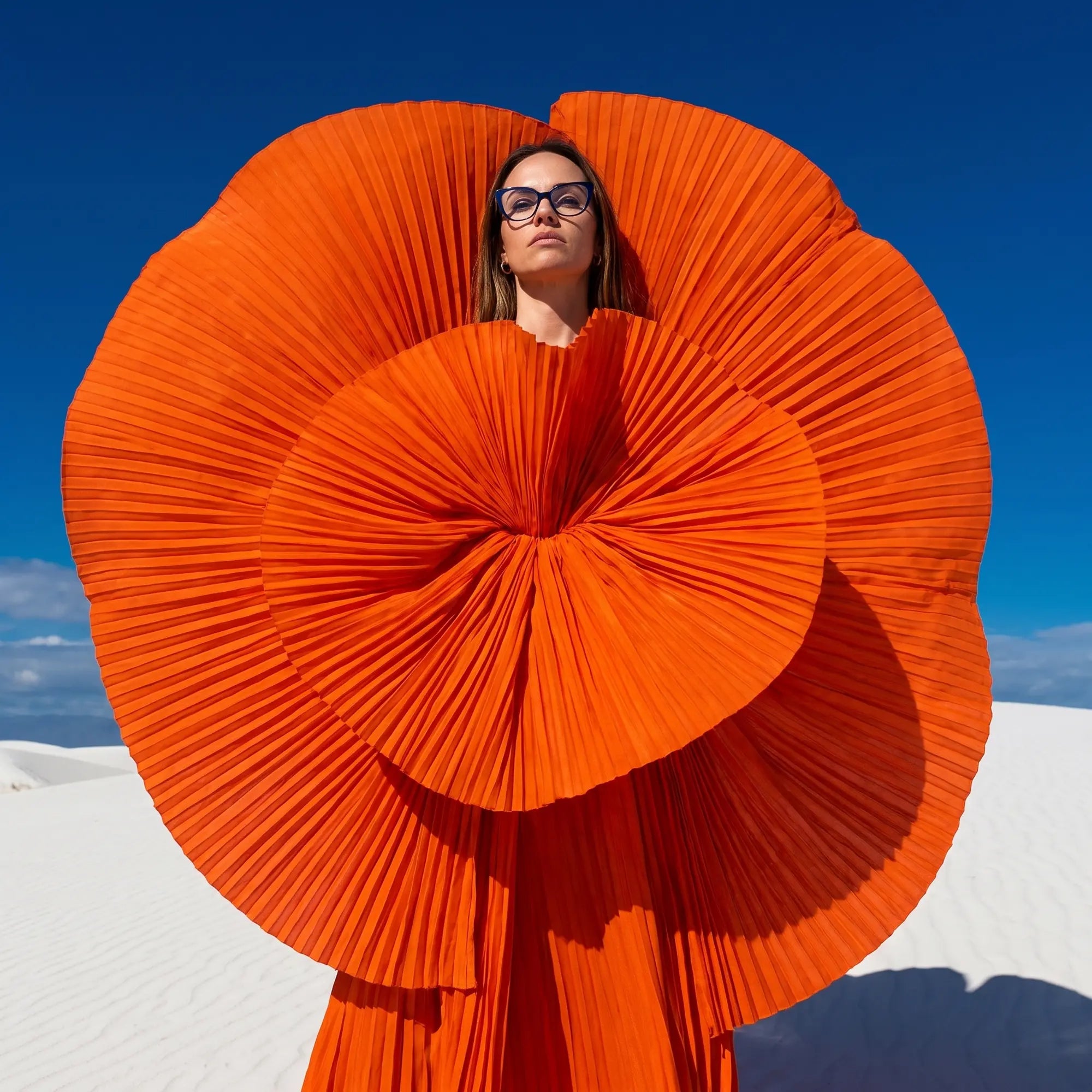 Fiore 1010 eyeglasses in blue, worn by a model – campaign portrait, sculptural orange pleated fan on white dunes