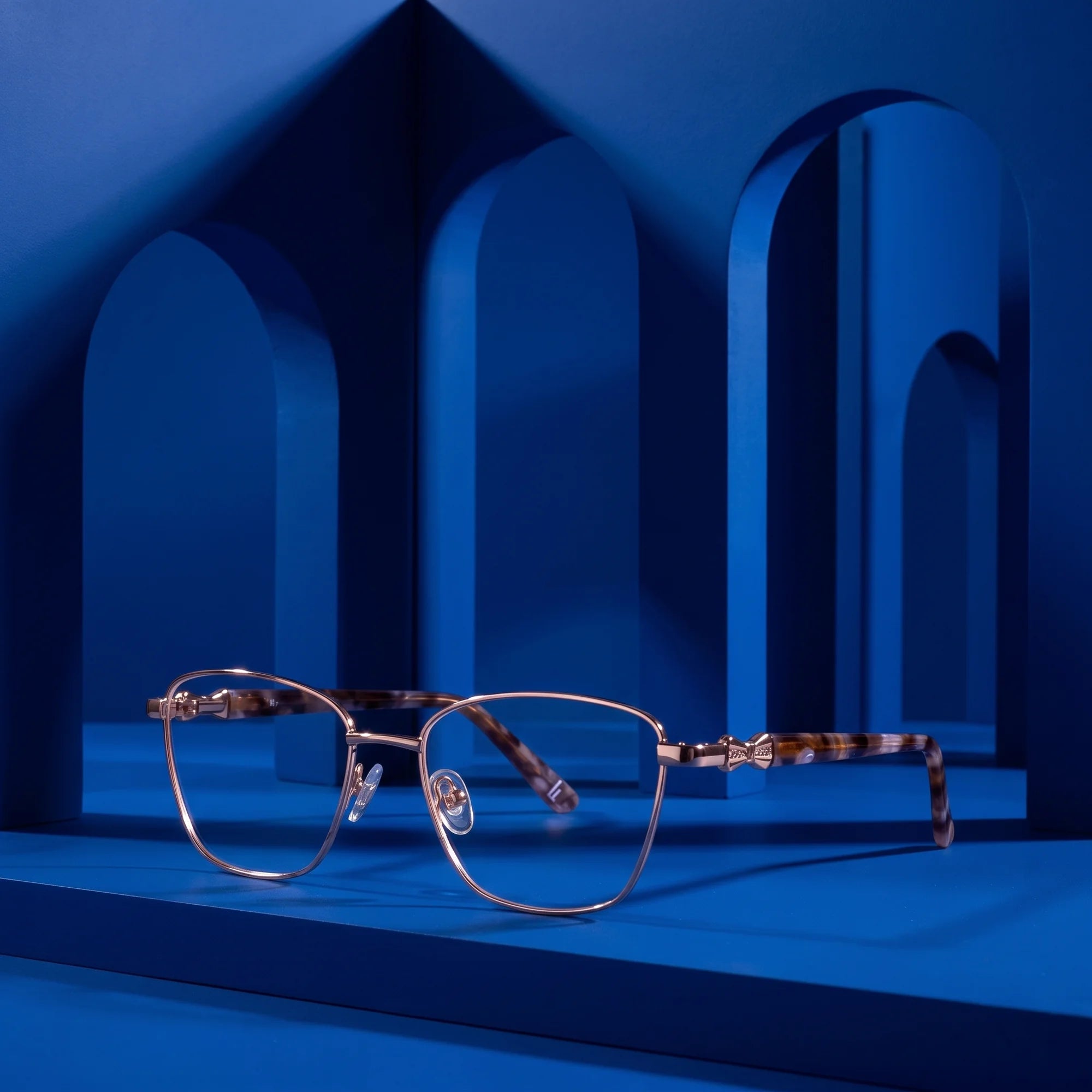 Fiore 22115 eyeglasses in gold – displayed on a cobalt podium among arches