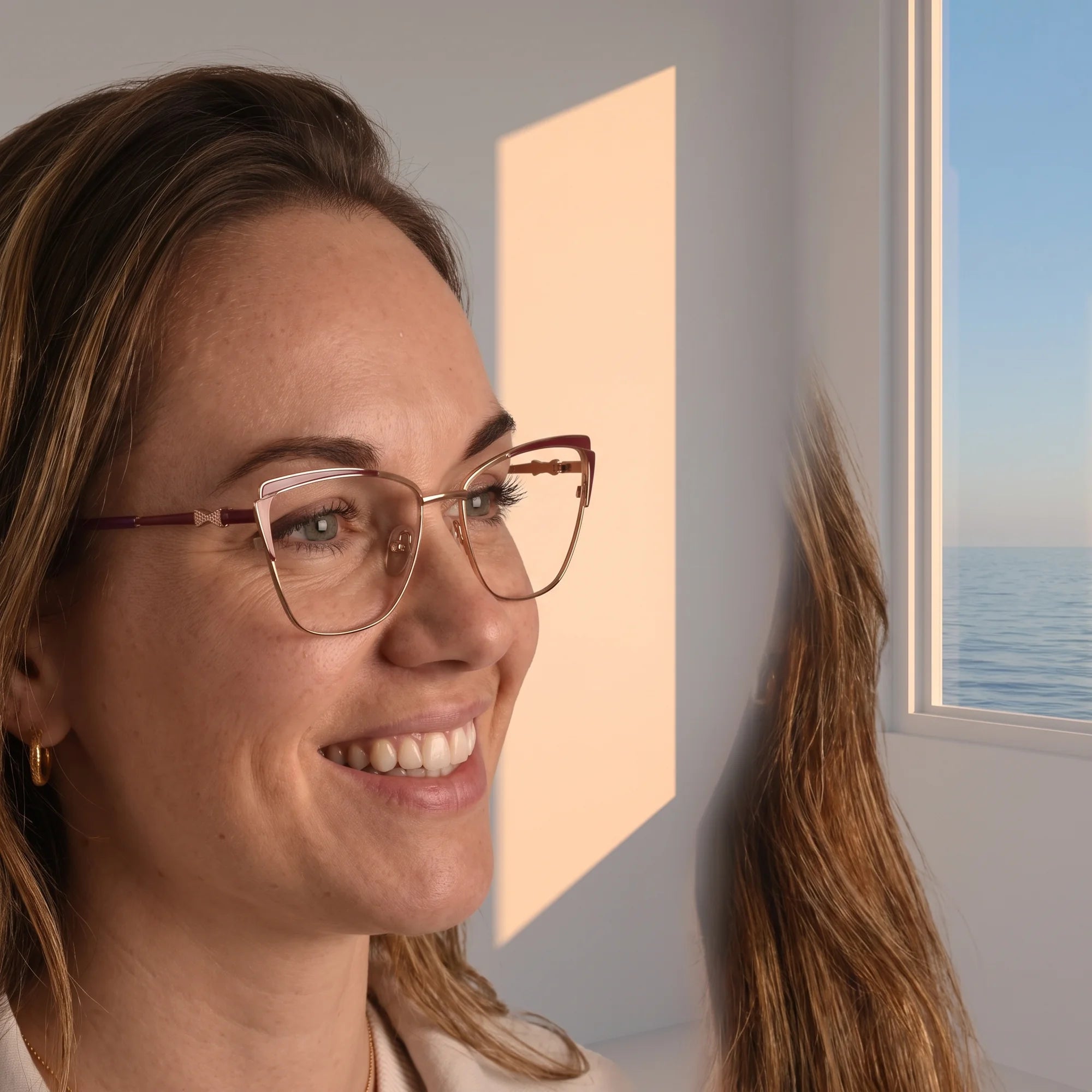 Fiore 3837 eyeglasses in red gold, worn by a model – campaign portrait, smiling at a bright window over the sea