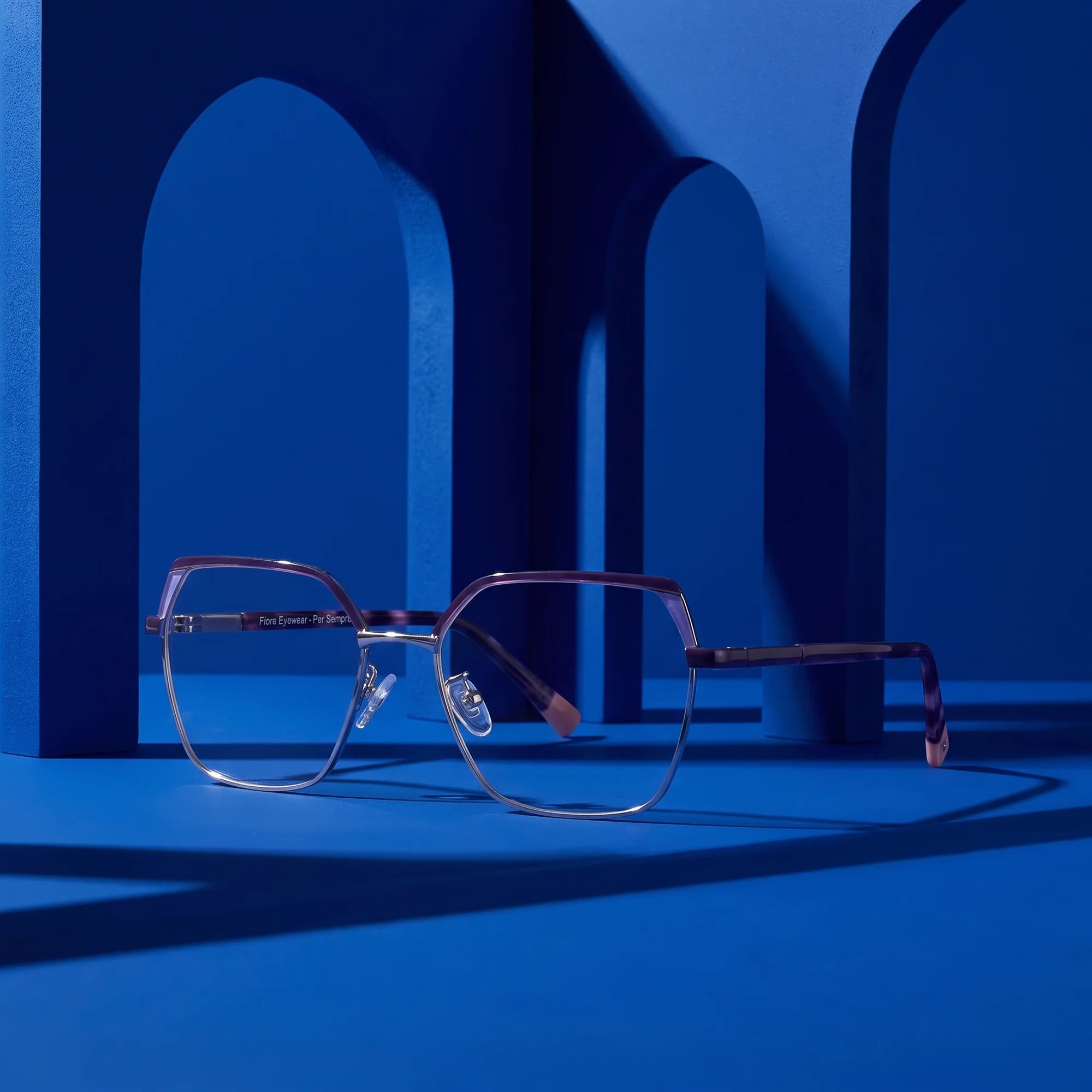 Fiore 3971 eyeglasses in purple silver – displayed on a cobalt podium among arches