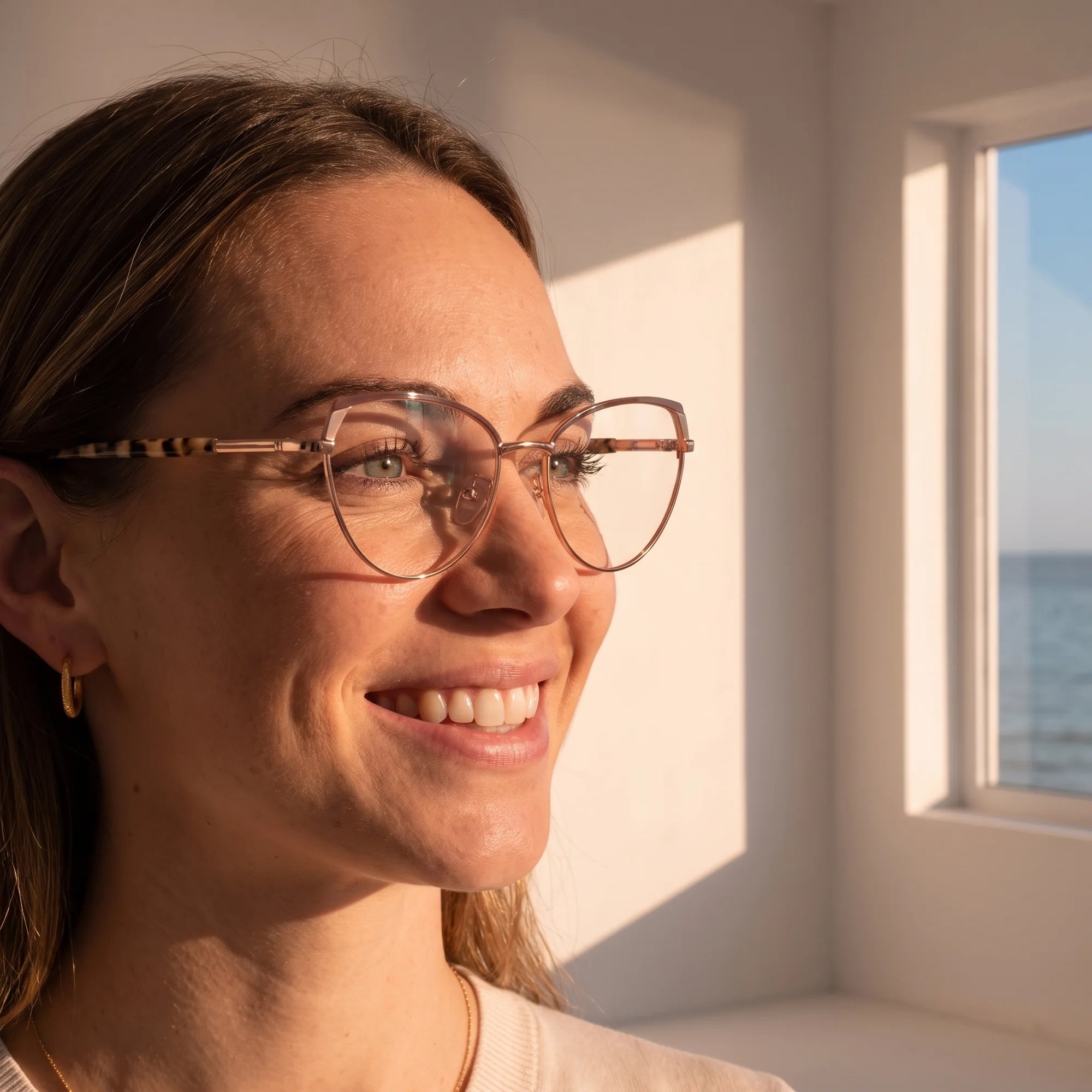 Fiore 3983 eyeglasses in brown gold, worn by a model – campaign portrait, smiling at a bright window over the sea
